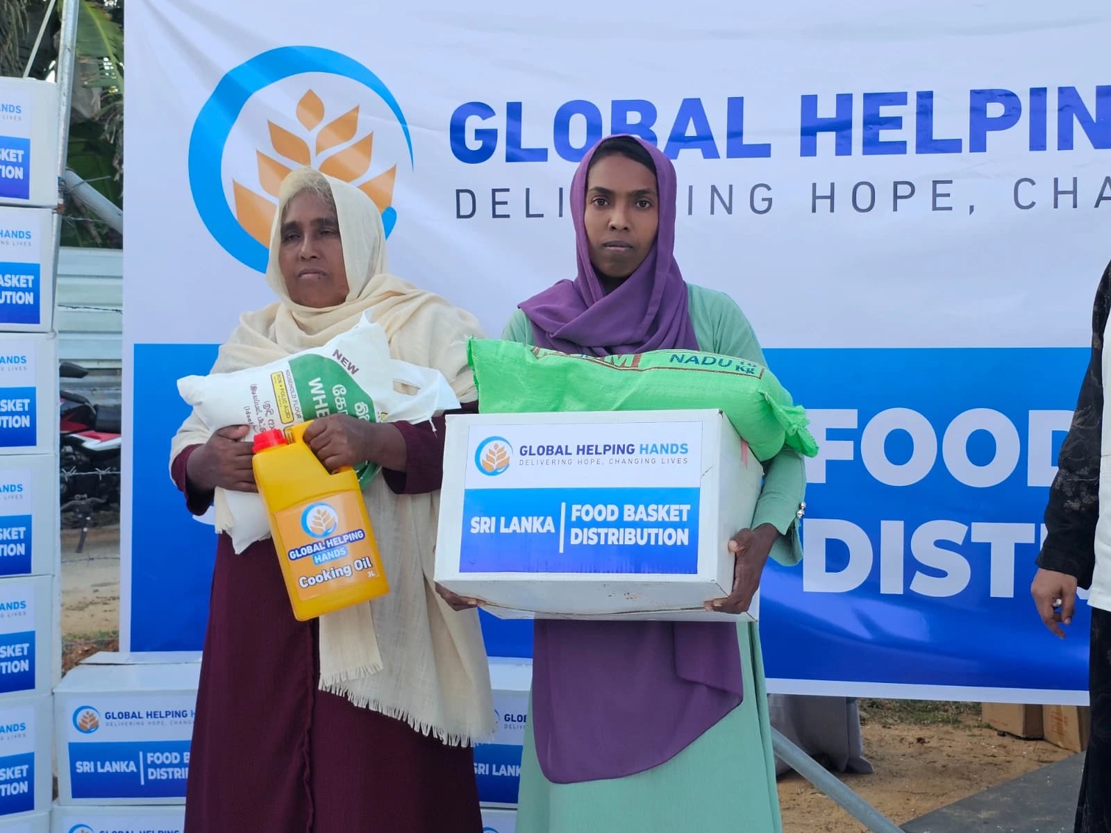 Two women holding a Global Helping Hands Sri Lanka food basket containing rice, wheat flour and cooking oil, in front of the partner banner