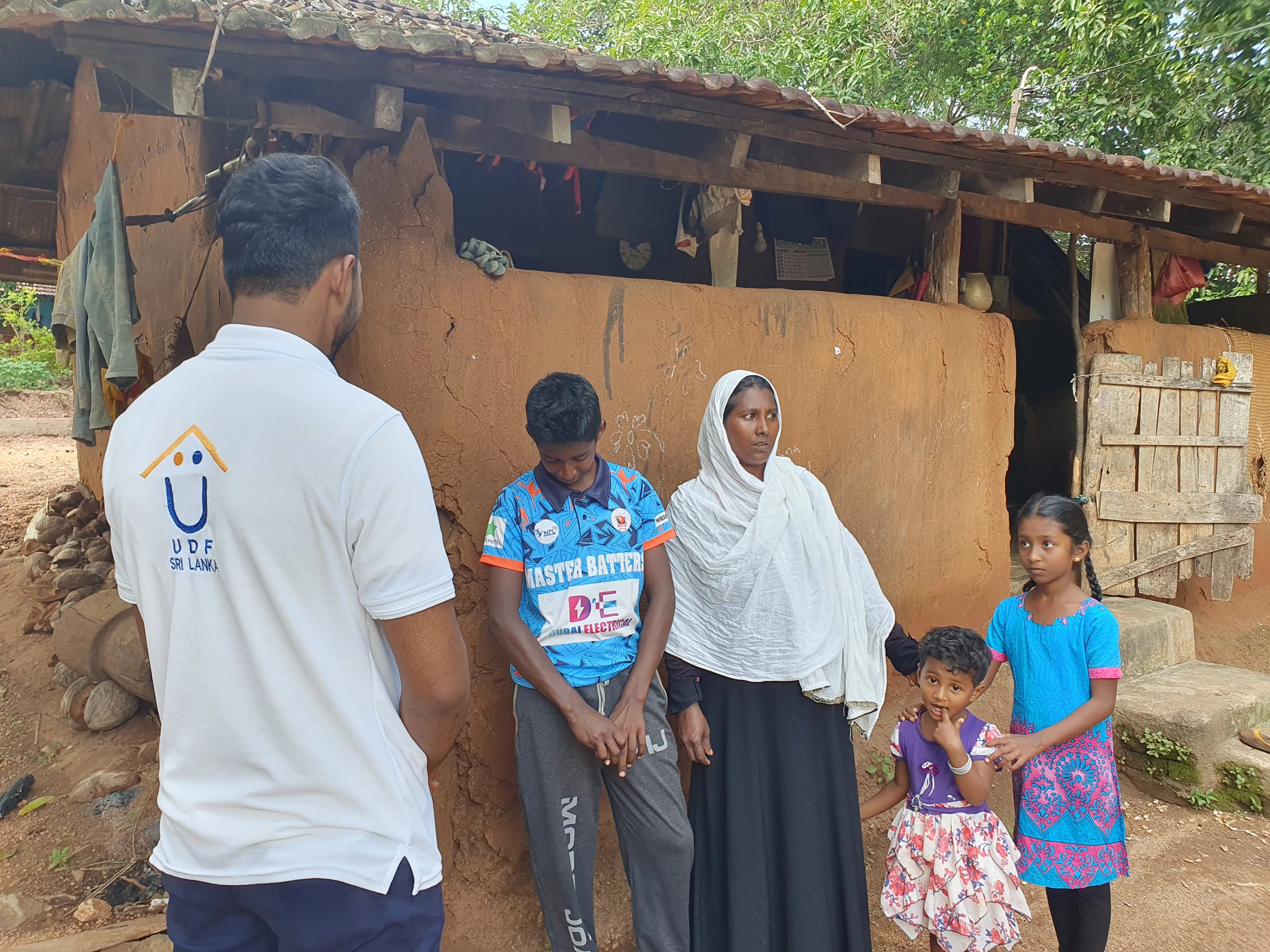 A UDF Sri Lanka field officer in a branded polo shirt speaking with a mother and her three children outside their mud-walled, tile-roofed home