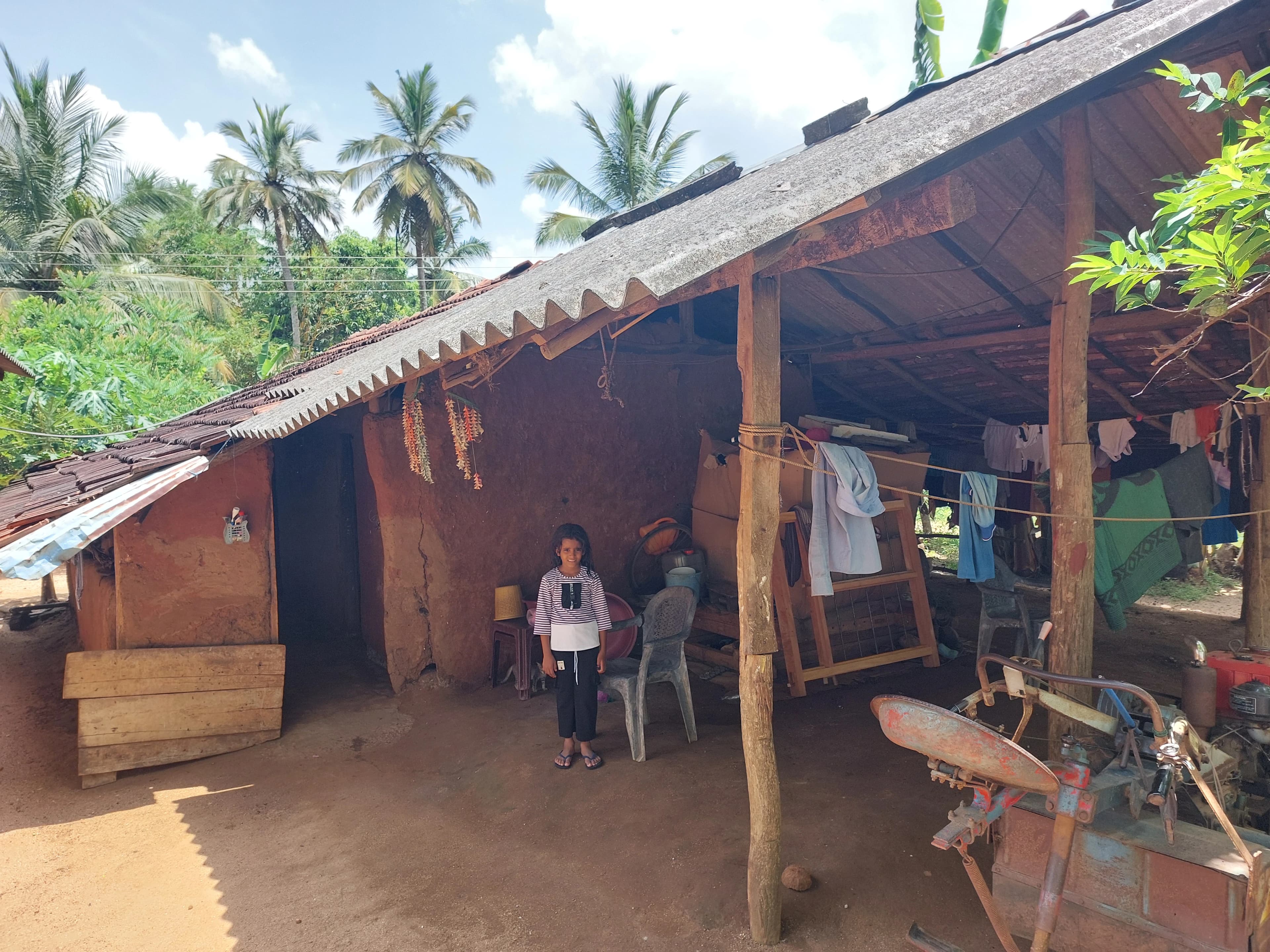 A child standing in front of a mud-walled village home with a corrugated roof, surrounded by coconut palms
